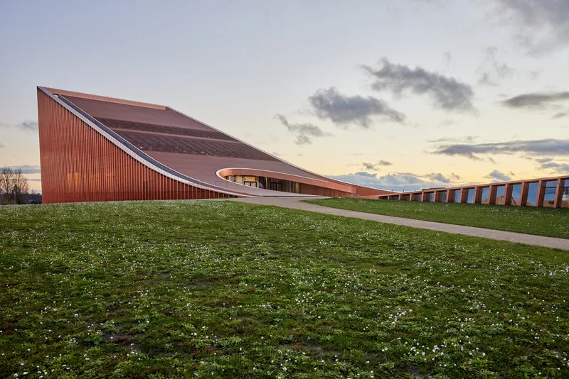 Sweeping terracotta roof lifting from the lawn at sunset with a curved colonnade in the distance