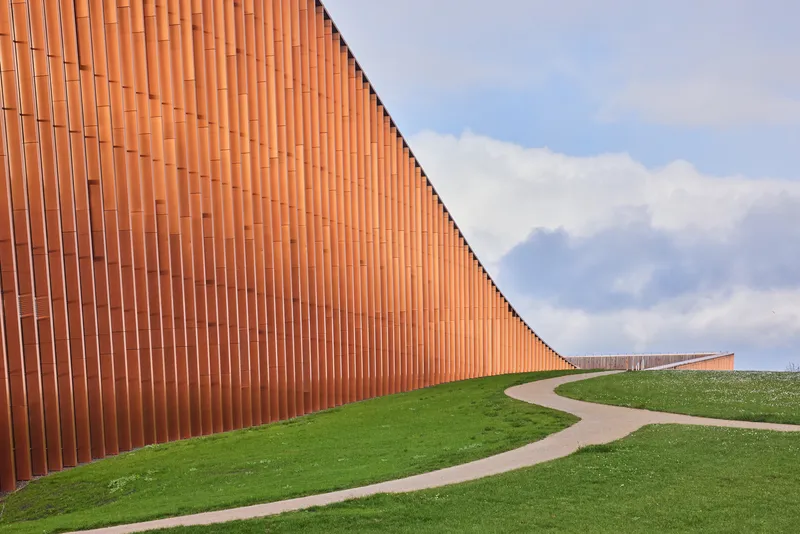 Curved path alongside the vertical terracotta cladding rising out of the grass under cloudy skies