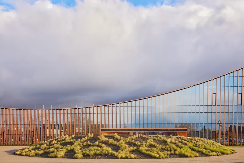 Curving timber and corten screen facade behind a circular planted bed under stormy clouds