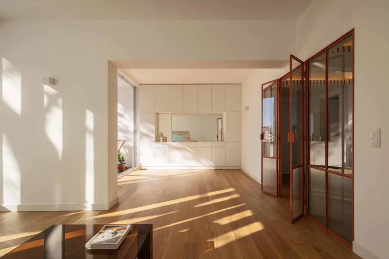Interior view through red-framed glazed doors toward a sunlit living space with white storage wall