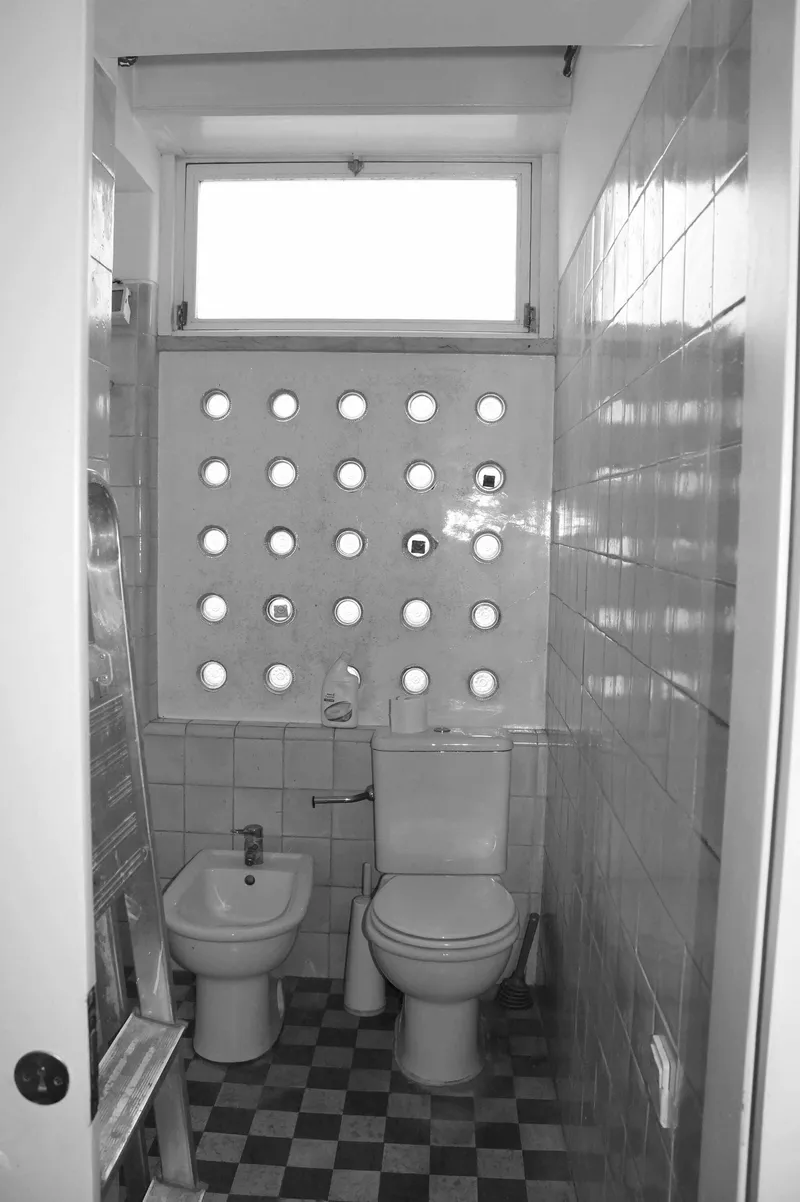 Narrow bathroom with circular perforated panel above toilet and bidet flanked by white tile walls