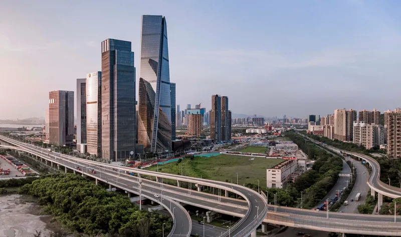 Cluster of glass towers framed by elevated highway ramps and green parkland at dusk