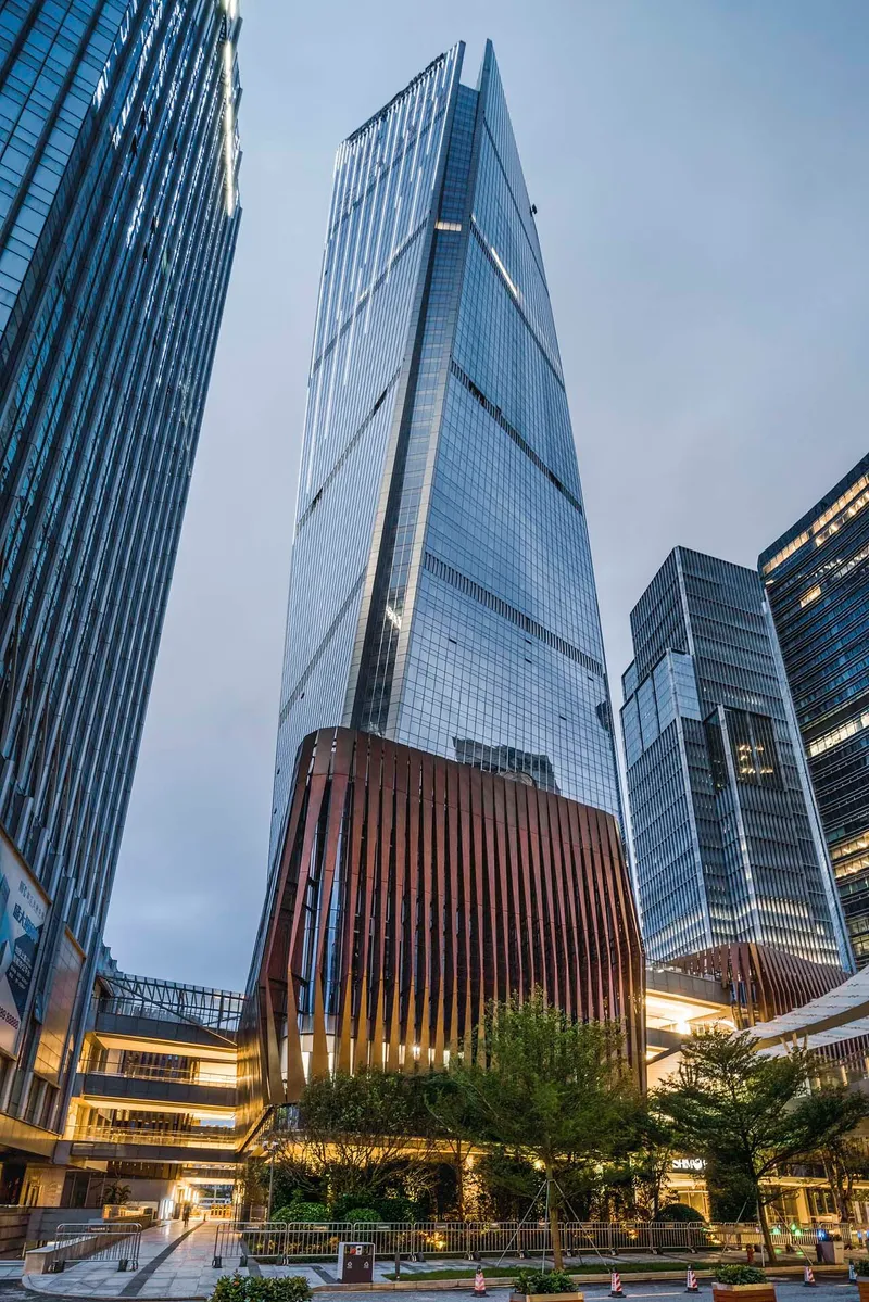 Upward view of the faceted glass tower with illuminated timber-clad base volume at twilight
