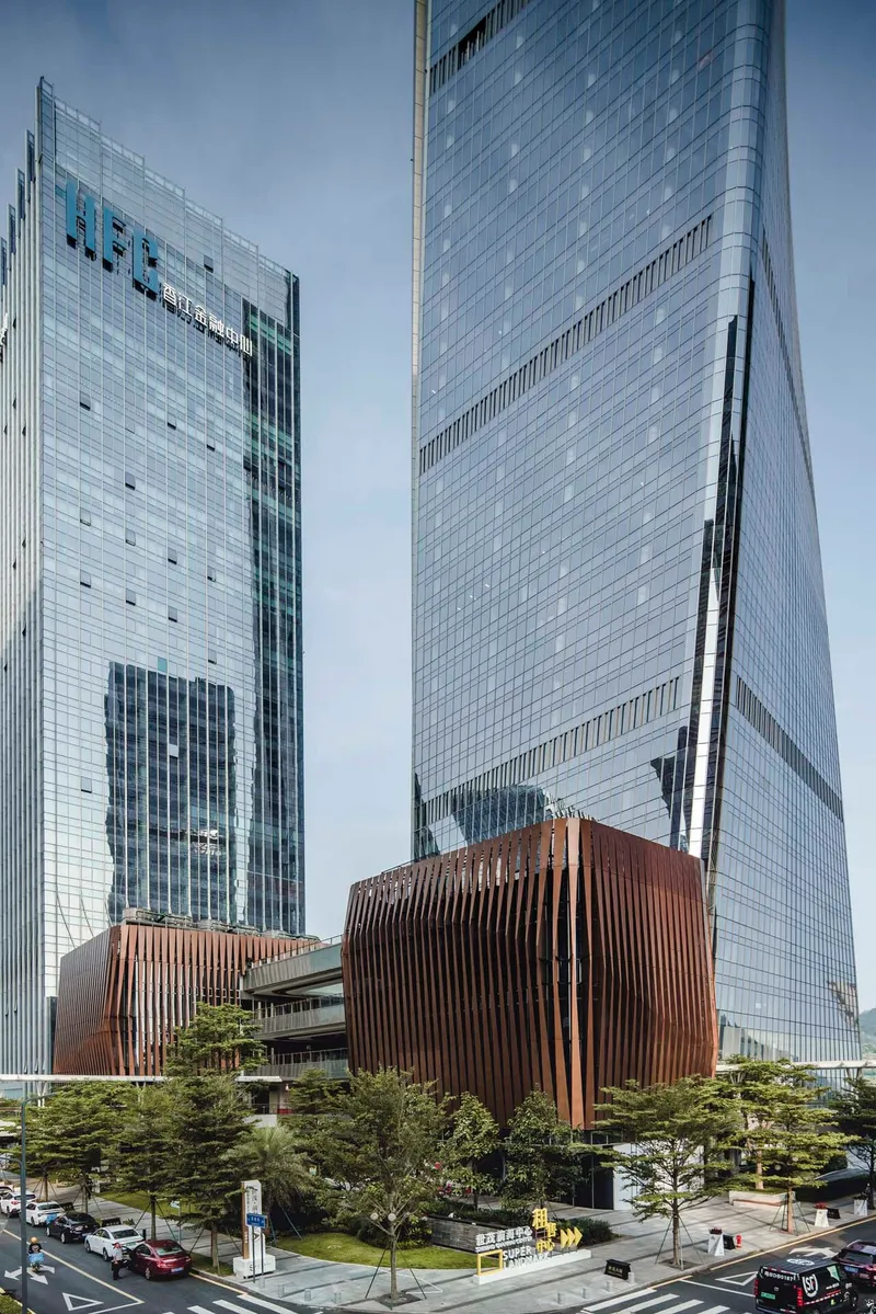 Curved bronze louvered pavilion at the base of the glass tower surrounded by planted evergreens