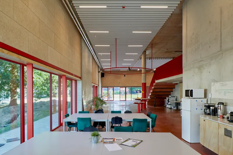 Double-height dining space with red steel staircase, plywood walls, and glazed doors opening to landscaped grounds
