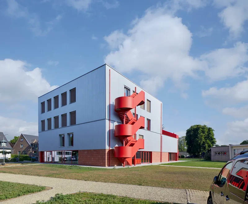 Street view showing the corrugated metal facade with a sculptural red stair tower against a partly cloudy sky