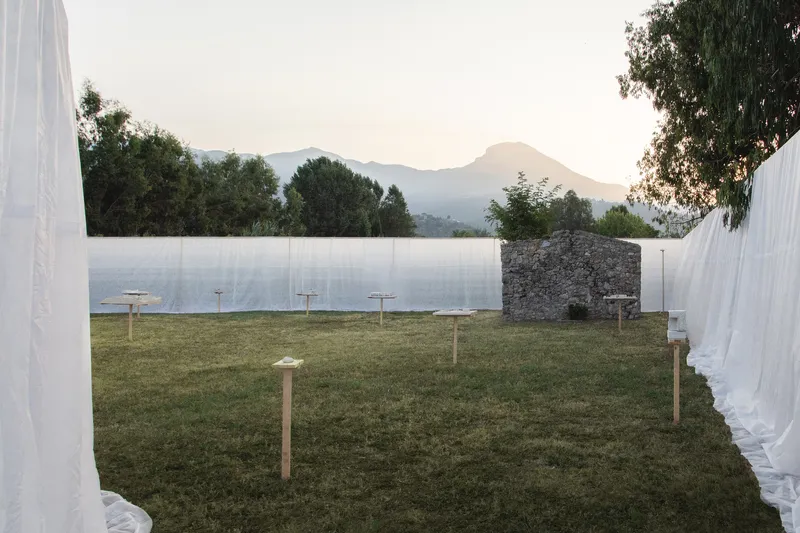Grass courtyard framed by white fabric curtains with a stone wall and distant mountains at dusk