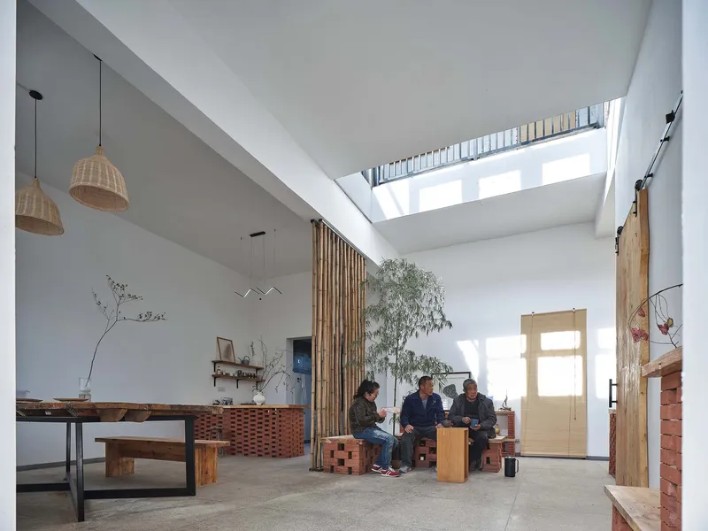 Interior living space with three people seated on timber stools beneath continuous skylight and bamboo screen