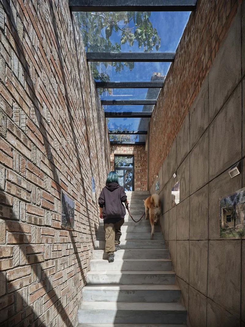 Narrow stairway between brick and concrete walls topped with steel beams and glass as two people ascend with a dog