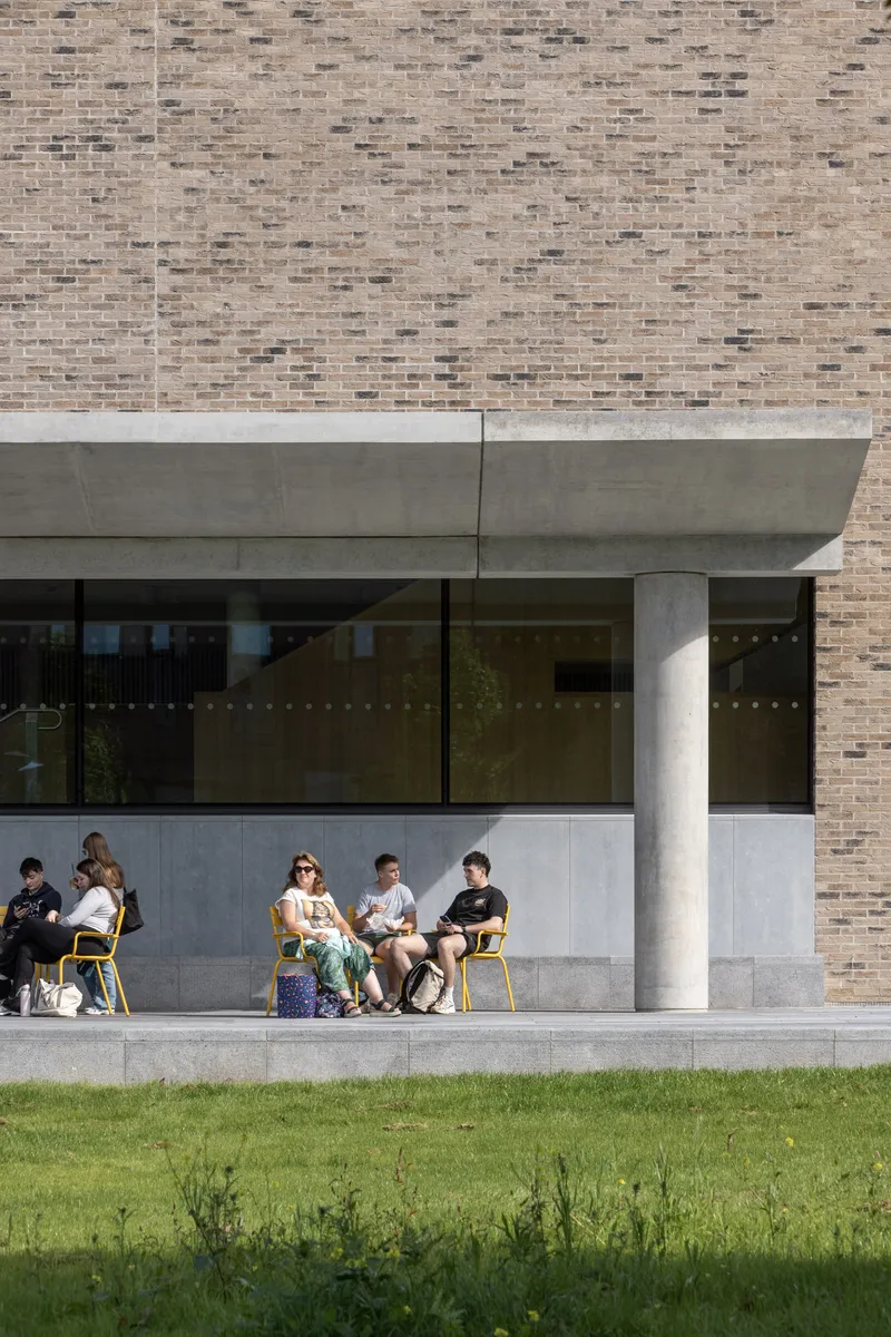 Covered terrace with concrete columns and beam supporting brick wall above students seated on yellow chairs