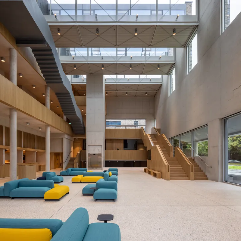 Multi-story atrium with timber clad balconies, concrete walls, and modular blue and yellow seating below clerestory windows