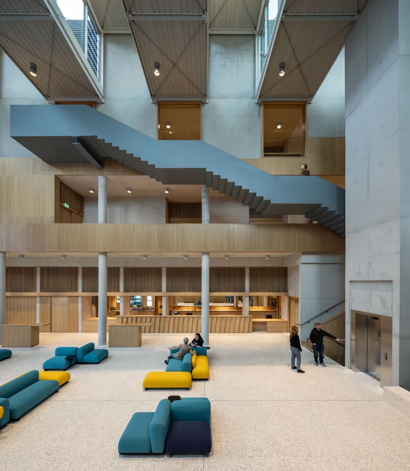 Cantilevered steel staircase crossing the atrium with timber-lined walls and visitors gathered on modular seating below