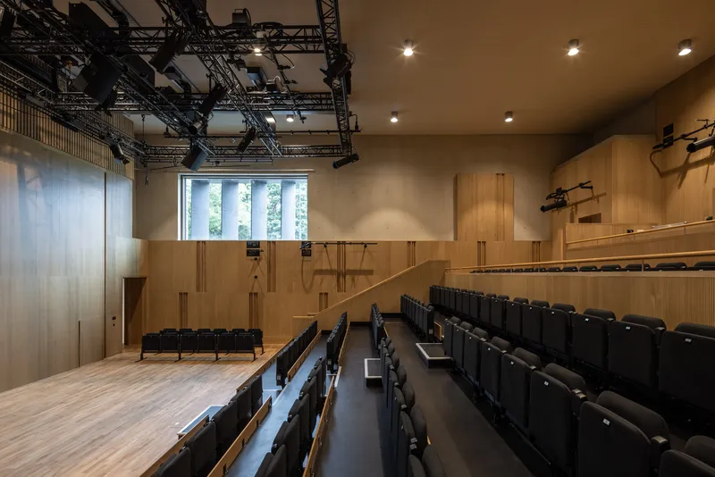 Auditorium interior with raked timber seating, exposed lighting truss, and vertical slatted wood walls with tall windows
