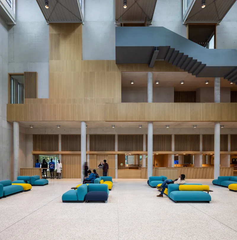 Lobby floor with timber service counter, concrete columns, and visitors seated on modular teal and yellow upholstered modules