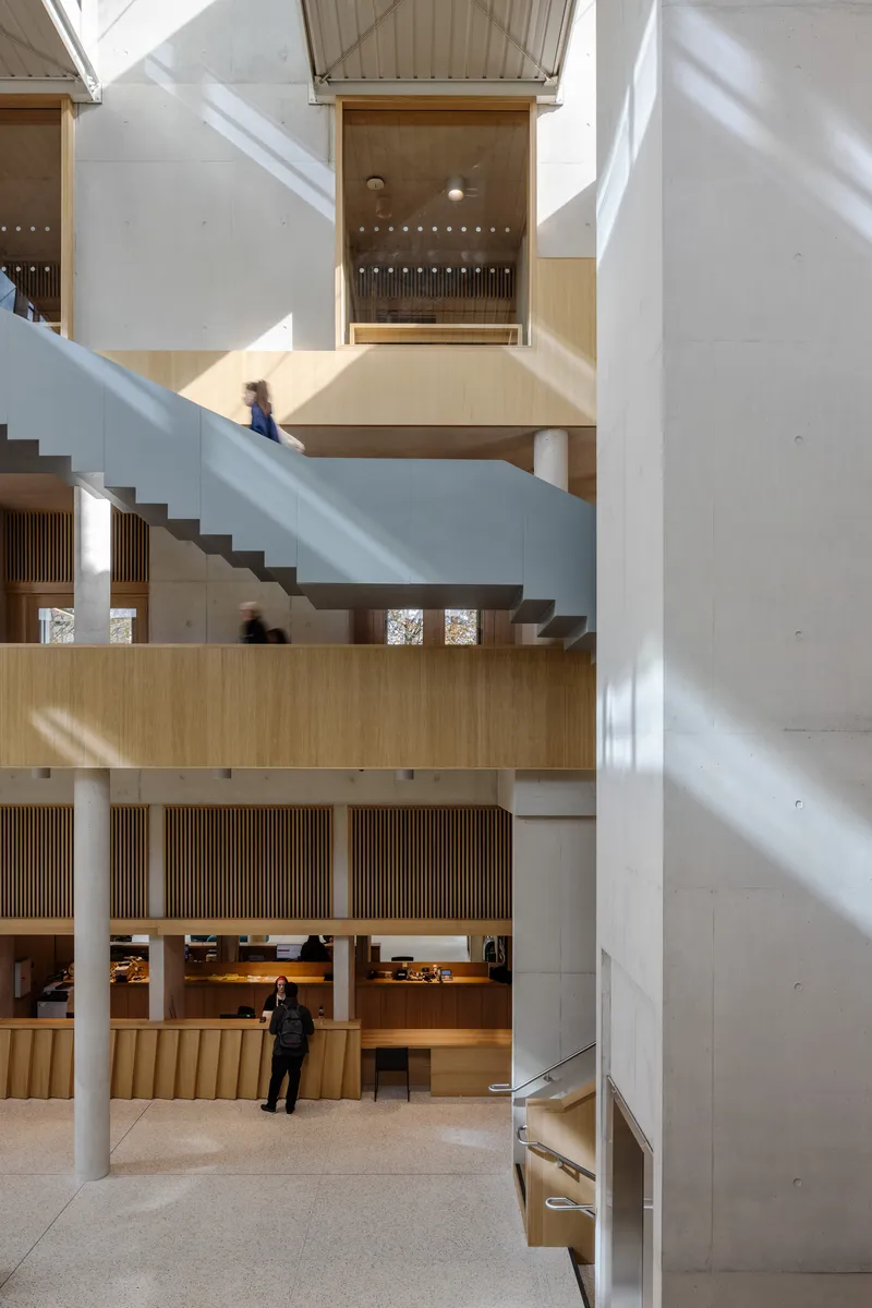 Multi-level atrium with concrete columns, timber-lined balconies, and a white staircase with figure below