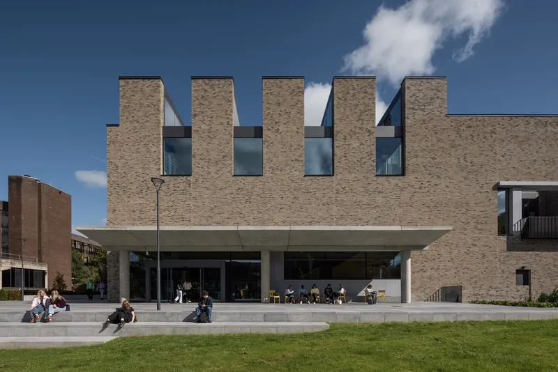 Brick facade with four vertical window slots and a recessed concrete canopy over students on terraced steps