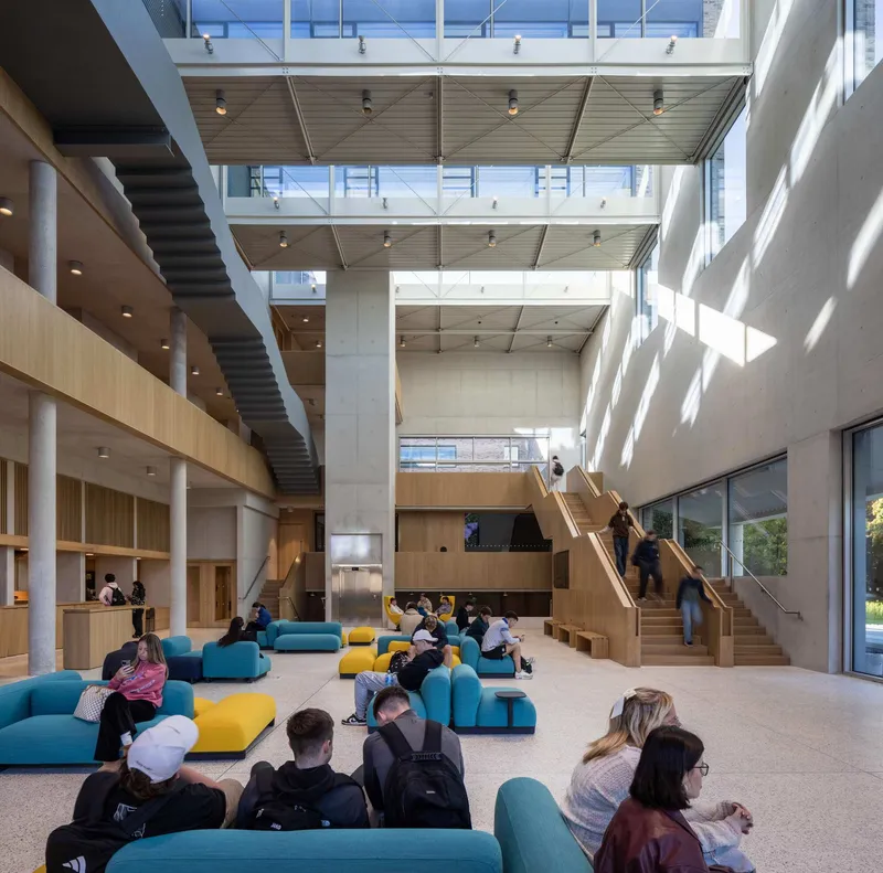 Triple-height atrium with timber ceiling panels, suspended bridges and students gathered on colorful floor cushions