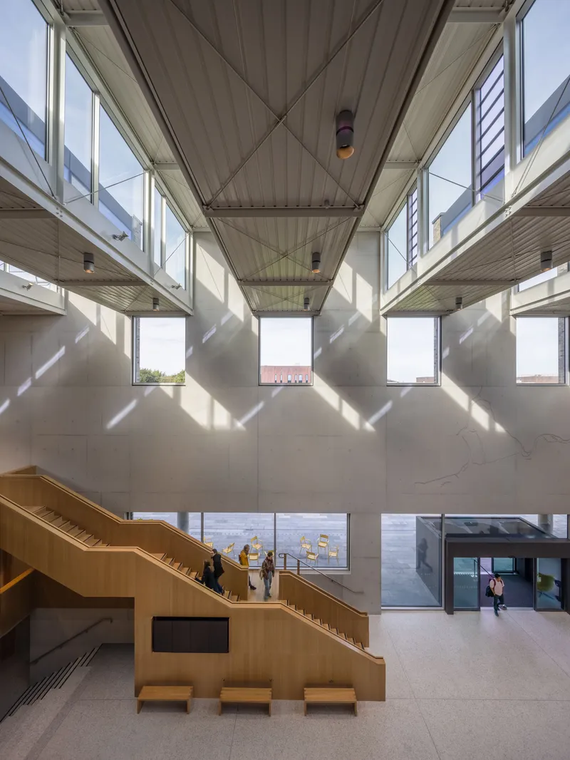 Four-story atrium looking down at timber staircase with diagonal light crossing suspended walkways and clerestory windows
