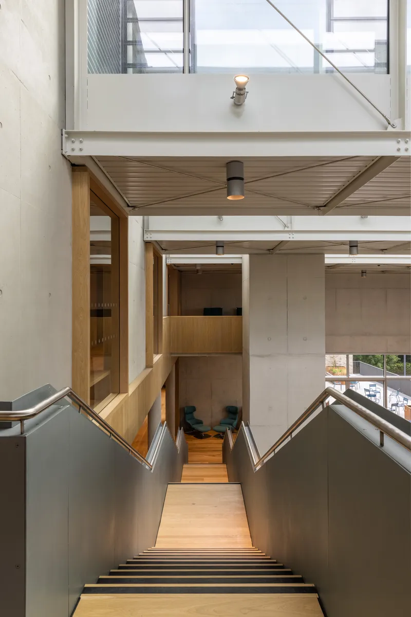 Staircase descending between metal walls toward timber landing with overhead skylights