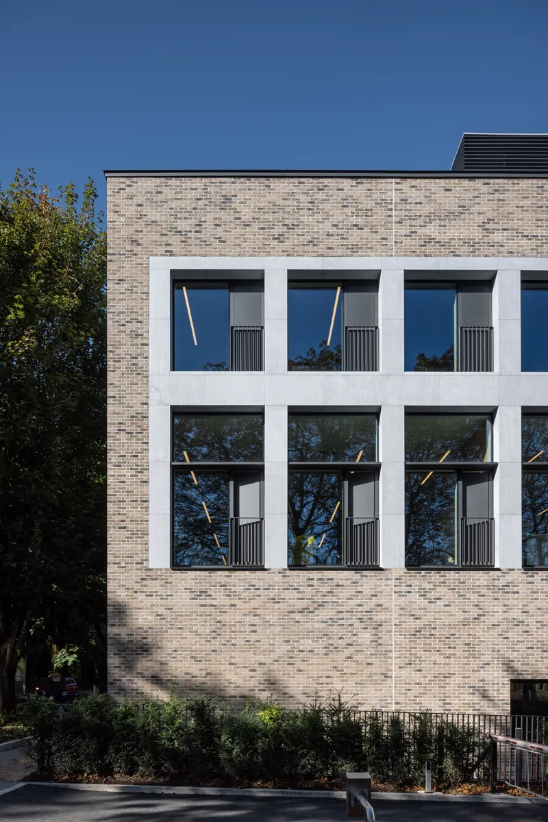 Buff brick facade with deep white window frames and trees reflected in glass under clear sky
