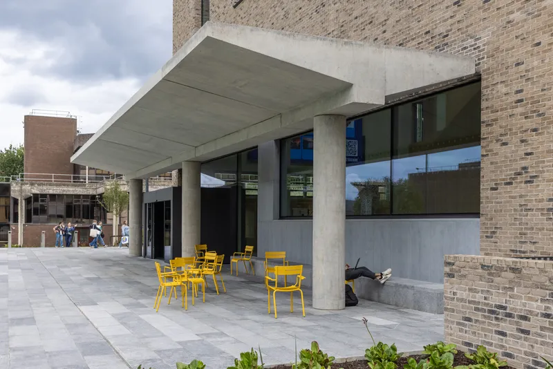 Covered concrete canopy sheltering yellow cafe chairs on paving beside brick wall