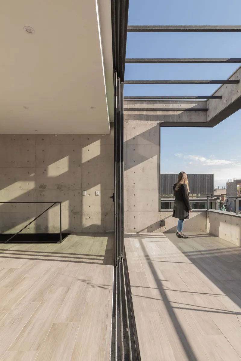 Sliding glass door opening to a concrete rooftop terrace with steel beam pergola and a person standing outside