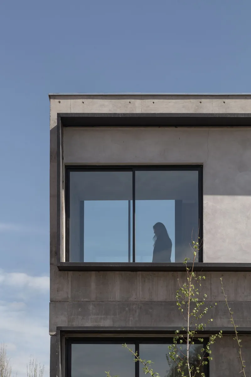 Upper level corner detail showing board-formed concrete facade with figure silhouetted behind glazing at dusk