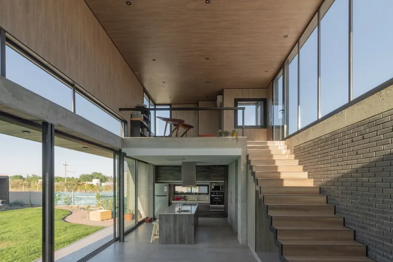 Double-height interior space with timber-clad mezzanine overlooking kitchen and timber stair against brick wall