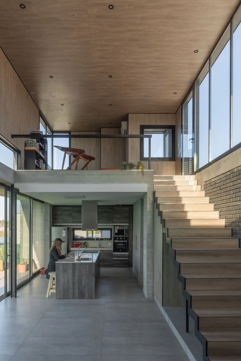 Double-height interior showing the wood-lined staircase ascending past the kitchen with a person seated below