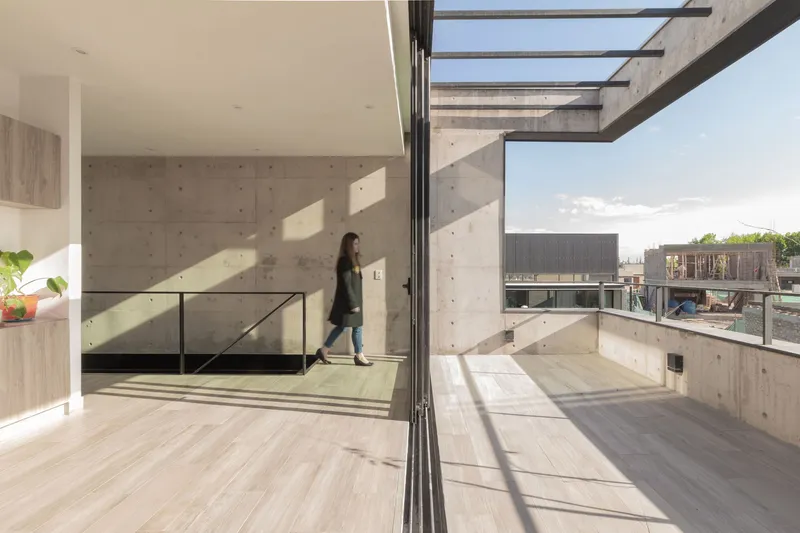 Upper-level terrace with concrete walls and glass railing where a person walks under the skylight opening