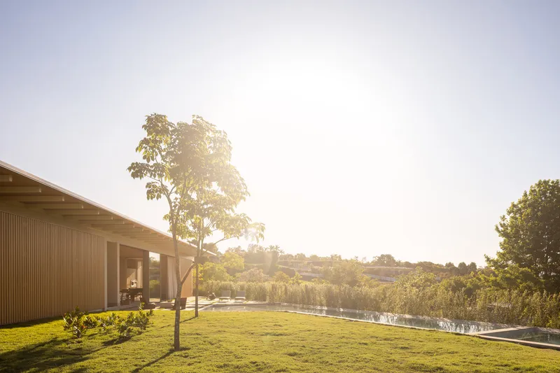 Single-story pavilion with timber cladding and overhanging roof beside a lawn and pond in warm afternoon light