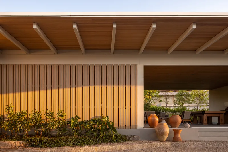 Covered porch with exposed timber rafters, vertical wood slat screen, and ceramic vessels beside planted beds at dusk