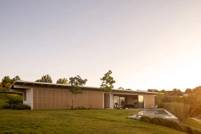 Long low-slung volume with horizontal wood cladding and projecting roof set into a sloping lawn at sunset