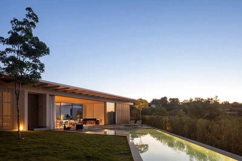 Timber-clad pavilion with overhanging roof and glazed walls beside a lap pool at dusk