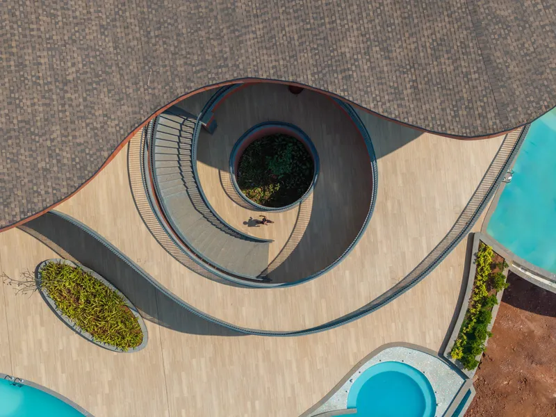 Aerial view of the curving concrete deck with a circular water feature and planted beds beside two pools
