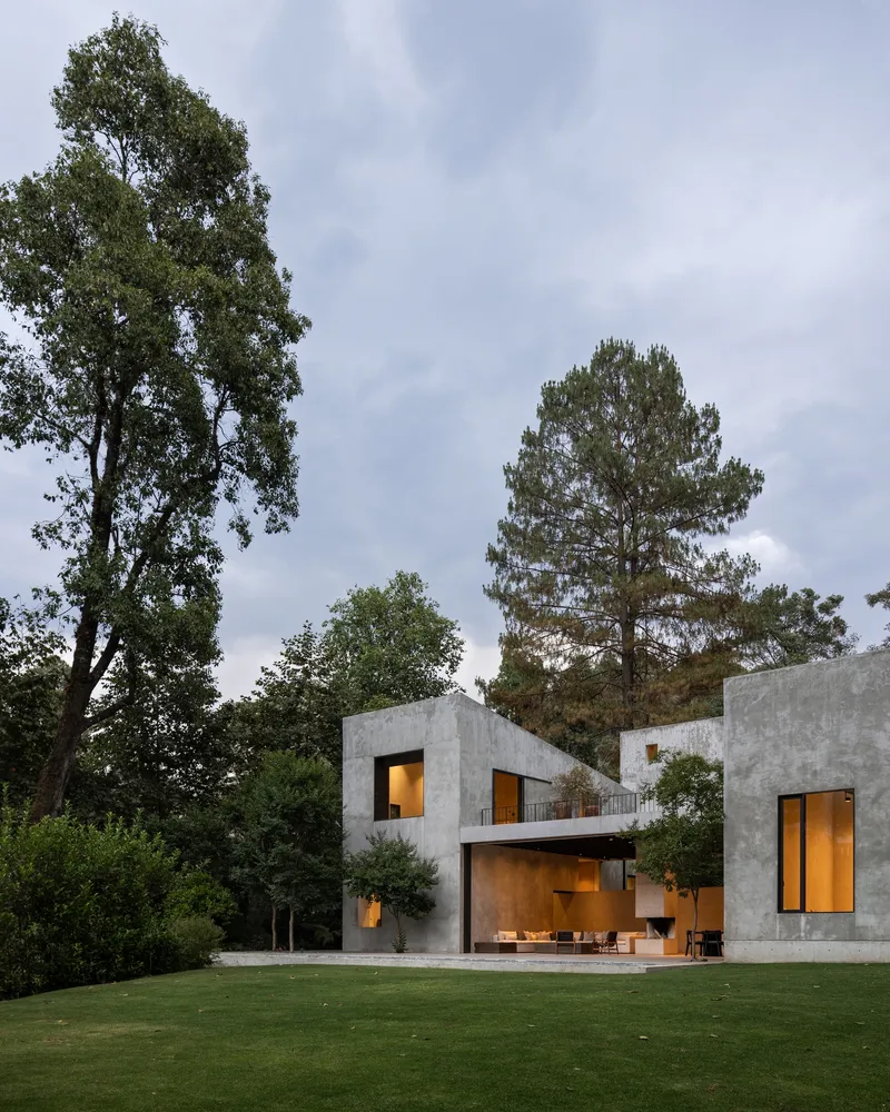 Concrete facade with illuminated openings at dusk beneath cloudy skies and surrounding forest