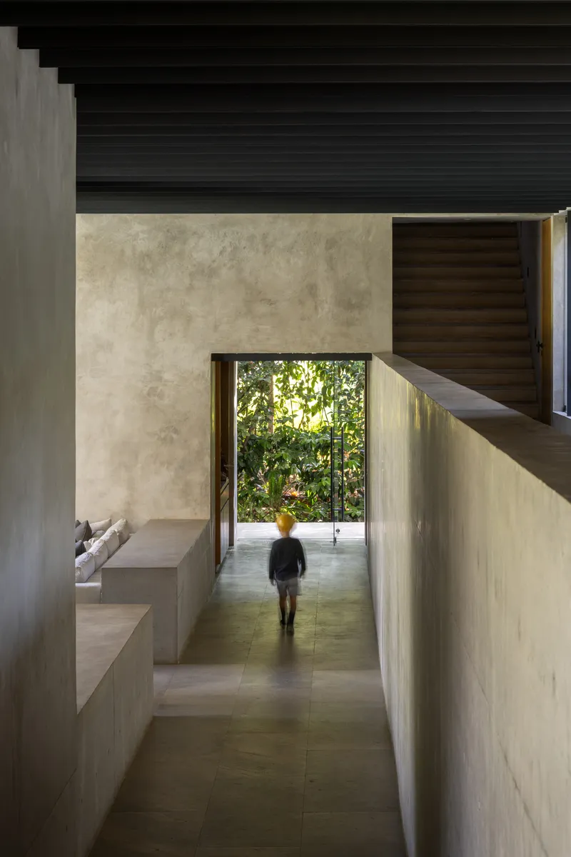 Entry corridor with figure walking toward garden framed by concrete walls and dark timber ceiling