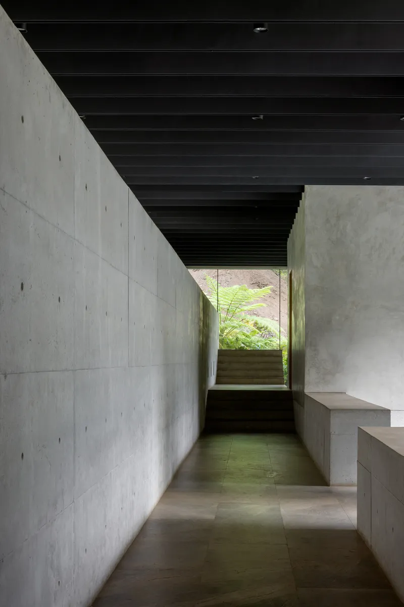 Concrete corridor with black ceiling beams leading to a sunlit window framing greenery beyond