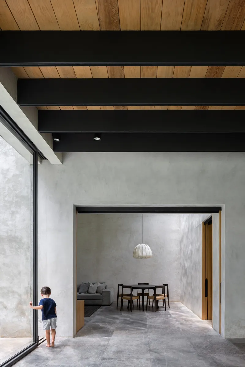 Living and dining space with exposed black beams and timber ceiling panels as a child stands at the glazed wall