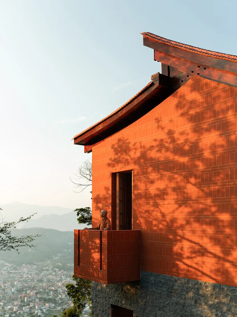 Cantilevered balcony with red tile cladding and figure standing at sunset with tree shadows cast across the facade