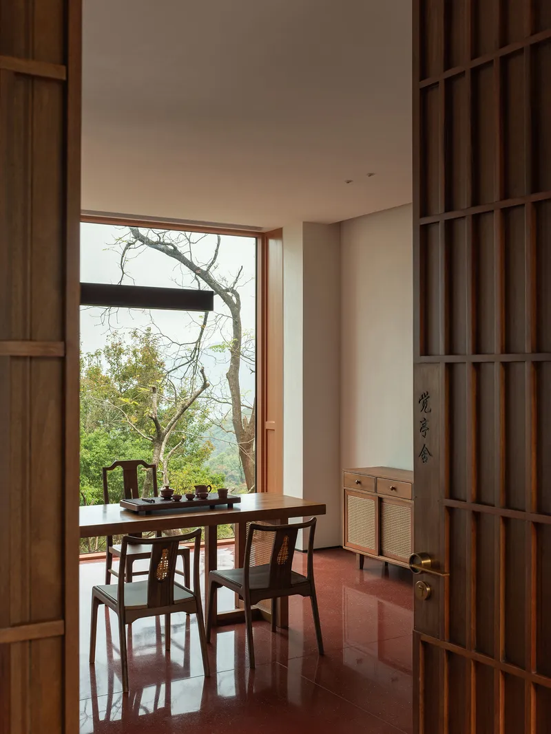 Dining space with timber table and chairs framed by open gridded doors facing a hillside tree