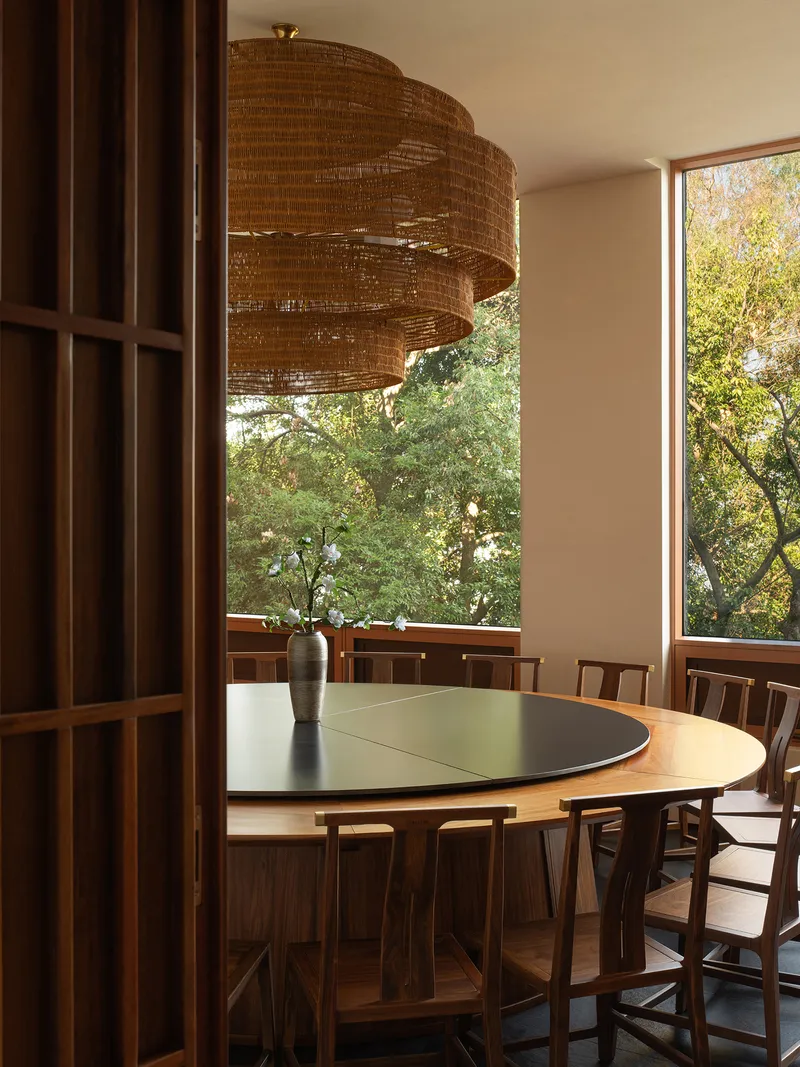 Round dining table beneath a woven pendant light with corner windows overlooking dense foliage