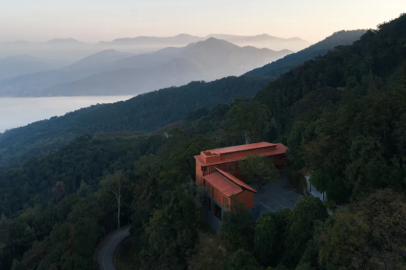 Aerial view of the angled red roof volumes nestled into forested hillside at dusk