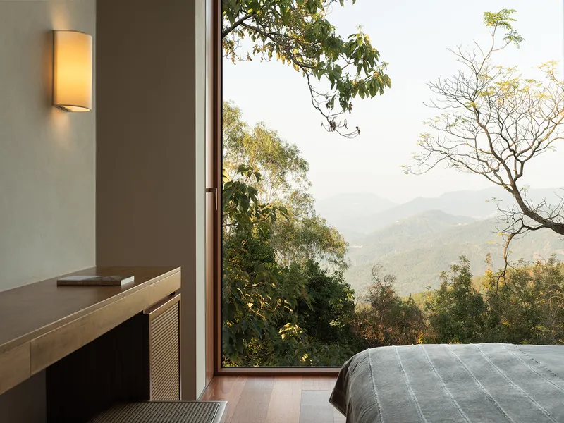 Floor-to-ceiling bedroom window overlooking forested hillside and distant mountains in daylight