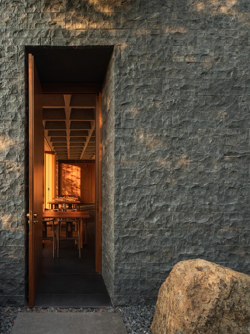Narrow entrance framed by stacked dark stone walls with warm interior lighting and a boulder beside gravel