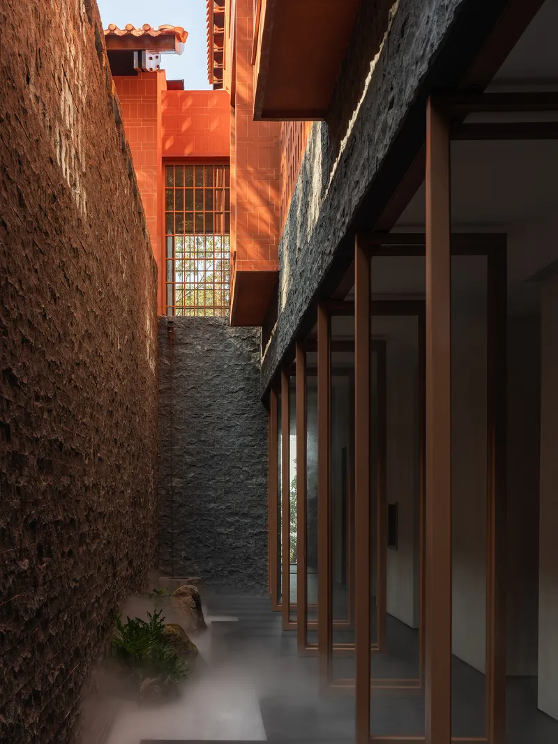 Courtyard passage between dark stone walls with timber screen and terracotta volumes under afternoon sun