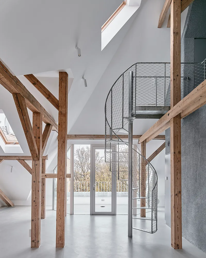 Metal spiral staircase with mesh railings rising through double-height space framed by timber posts and glazed doors