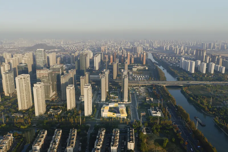Wide aerial showing the low-rise roof among dense residential towers and a tree-lined canal