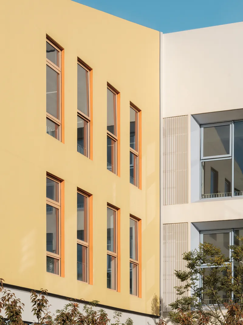 Close-up of the yellow facade with orange-framed windows meeting a white ribbed-panel volume and small tree