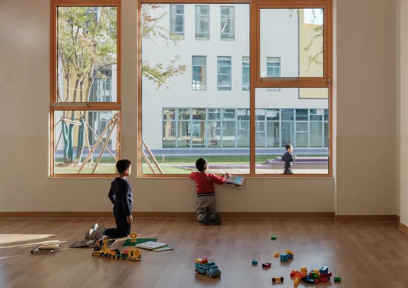 Interior classroom with timber-framed windows overlooking the courtyard where two children sit with scattered toys on the floor
